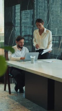 İş adamı elinde belgelerle gülen meslektaşı yanında dururken bir dizüstü bilgisayarda çalışıyor. Mermer masası ve pencereleri olan parlak ve modern bir ofiste iş birliği yapıyorlar..