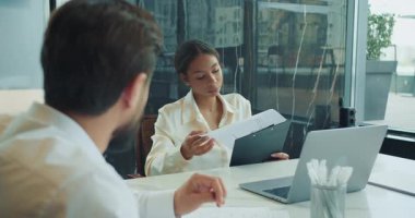 İş kadını, modern bir ofisteki bir toplantıda erkek meslektaşına verileri panoda sunar. Laptop ve evraklar beyaz masada görünüyor. Yüksek kalite 4k görüntü