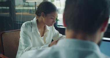 Genç iş kadını profesyonel bir toplantı sırasında basılı belgeleri dikkatle inceliyor. Erkek arkadaşı masanın karşısında oturuyor. Parlak, modern ofis ortamında, doğal ışık altında. Yüksek