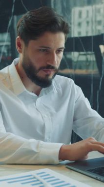 Beyaz gömlekli sakallı bir iş adamı modern bir ofiste dizüstü bilgisayarla çalışıyor. Tablolar ve masadaki kahve fincanı veriye dayalı bir çalışma oturumu öneriyor..