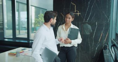 İş adamı ve iş kadını resmi giysiler içinde dizüstü bilgisayar ve belgeleri mermer duvarları ve parlak gün ışığı olan modern bir ofiste tutarken gündelik bir konuşma yapıyorlar. Yüksek kalite 4k görüntü