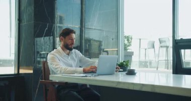 Modern bir ofis masasında dizüstü bilgisayarda çalışan resmi beyaz gömlekli bir iş adamı. Etrafı büyük pencereler ve doğal gün ışığıyla çevrili kahve ve belgelerle dolu. Yüksek kalite 4k görüntü