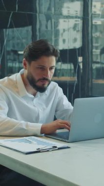 Modern bir ofis masasında dizüstü bilgisayar üzerinde çalışan beyaz gömlekli, odaklanmış iş adamı. Rapor, kahve ve akıllı telefon. Parlak gün ışığı geniş pencerelerden akar.