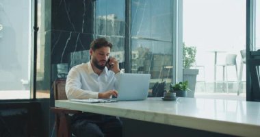 Beyaz gömlekli bir iş adamı, büyük pencereli, modern bir ofisteki bir masada otururken telefonla konuşarak ve dizüstü bilgisayarını işaret ederek birden fazla iş yapıyor. Yüksek kalite 4k görüntü