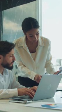 İşadamı dizüstü bilgisayarda yazarken bir iş kadını da yanındaki belgeyi yakından inceliyor. Cam duvarları ve yansımaları olan parlak bir ofis masasında birlikte çalışıyorlar..