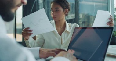 Beyaz gömlekli odaklanmış bir iş kadını modern bir ofiste yapılan bir tartışma sırasında kağıt bir belgeyi inceliyor. Erkek bir meslektaşım ön planda dizüstü bilgisayarda çalışıyor, görevler üzerinde işbirliği yapıyor. Yüksek kalite 4k