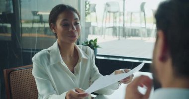 Beyaz bluzlu kendine güvenen bir iş kadını, modern, güneşli bir ofiste cam duvar ve açık hava manzaralı bir ofiste yaptığı toplantıda, yazılı bir belgeyi erkek arkadaşına verirken gülümsüyor. Yüksek kalite 4k