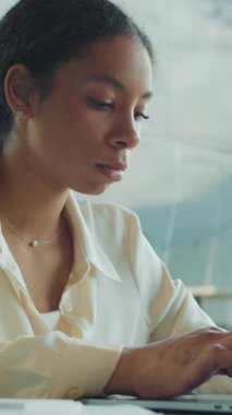 Odaklanmış genç iş kadını elinde kalemle masa başında oturuyor ve modern, parlak bir ofis ortamında çalışırken özenle dizüstü bilgisayarına bakıyor..