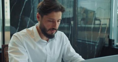 Beyaz gömlekli bir iş adamı modern bir ofiste dizüstü bilgisayarı üzerinde çalışırken konsantre olur. Arkasındaki camda yansımalarla pencerenin yanında oturuyor. Yüksek kalite 4k görüntü