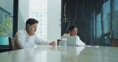 İş adamı telefonda notlar alırken konuşur ve bir iş kadını dizüstü bilgisayarın yanında belgeler okur. Pürüzsüz bir ofis ortamında büyük bir masada birlikte çalışıyorlar. Yüksek kalite 4k görüntü