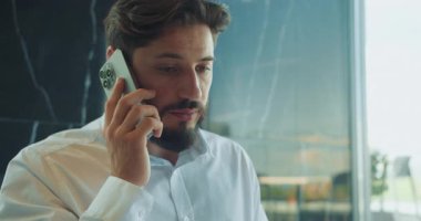 Beyaz gömlekli bir iş adamı kapalı alanda akıllı telefonla ciddi ciddi konuşuyor. Arka planda modern cam bir duvar var, profesyonel bir ofis ortamını işaret ediyor. Yüksek kalite 4k görüntü