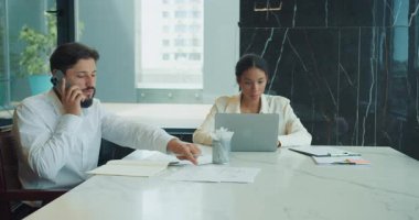 İş adamı telefonda konuşur ve bir iş kadını dizüstü bilgisayarda çalışırken notlar yazar. Her ikisi de modern ofis ortamında büyük bir masada odaklanmış ve çalışıyor. Yüksek kalite 4k görüntü