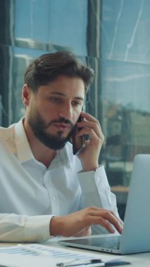 Beyaz gömlekli, kendine güveni tam bir işadamı telefonda konuşuyor ve modern bir ofis masasında, parlak bir dizüstü bilgisayarla kahve, belgeler ve doğal ışık ile yazı yazıyor..