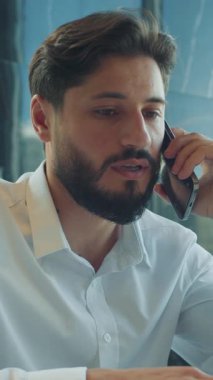 Modern bir ofiste dizüstü bilgisayar üzerinde çalışırken kendine güvenen bir iş adamı telefon görüşmesi yapıyor. Doğal ışık ve çağdaş iç mekan ile profesyonel ortam.