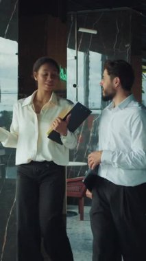 İki iş adamı, bir erkek ve bir kadın, modern bir ofis koridorunda el ele yürüyorlar, panoları tutuyorlar ve cam ve yeşillikle çevrili odaklanmış bir sohbete giriyorlar..