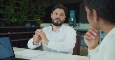 İşadamı basılı bir tablo üzerinde çalışıyor ve modern bir ofiste yapılan bir toplantıda bir meslektaşına verileri açıklıyor. Çizgi grafiklerini gösteren bir dizüstü bilgisayar ekranı da görünür. Yüksek kalite 4k görüntü