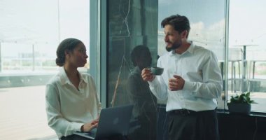 Modern bir ofiste iki profesyonel sıradan bir konuşma yapıyor. Kadın dizüstü bilgisayarla otururken, erkek kahve fincanı tutuyor. Bu da ortak çalışma ortamını yansıtıyor. Yüksek kalite
