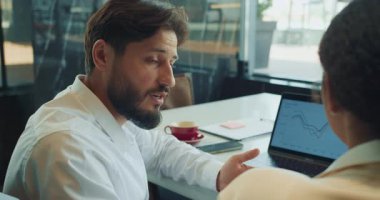 Beyaz gömlekli bir işadamı, modern ofis ortamında yapılan bir toplantıda dizüstü bilgisayarda gösterilen renkli bir çizelgeye işaret ederek, veri eğilimlerini bir meslektaşına anlatıyor. Yüksek kalite 4k görüntü