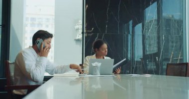 İş adamı telefonda konuşur ve notlar yazar. Bu sırada bir iş kadını dizüstü bilgisayarın yanındaki panoya bakar. Belgeler ve grafiklerle dolu modern bir ofis masasında oturuyorlar. Yüksek kalite 4k görüntü