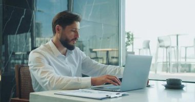 Temiz, modern bir ofis masasında dizüstü bilgisayar üzerinde çalışan beyaz gömlekli, odaklanmış işadamı büyük camlardan dökümanlar, kahve ve doğal gün ışığı akıyor. Yüksek kalite 4k görüntü