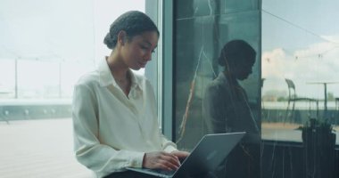 Modern bir ofiste pencerenin yanında oturan genç bir iş kadını dizüstü bilgisayarla çalışıyor. Mermer duvardaki yansıması, temiz ve profesyonel atmosfere derinlik katıyor. Yüksek kalite 4k görüntü