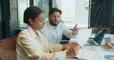 İki meslektaş, bir erkek ve bir kadın, parlak ve modern bir ofiste finansal bir belgeyi tartışıyorlar. Dosya, dizüstü bilgisayar ve not defterleriyle birlikte veri analizine odaklanmış bir masada oturuyorlar. Yüksek kalite 4k