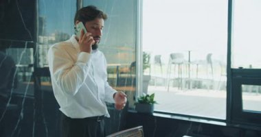 Beyaz gömlekli bir işadamı, bir elini sandalyeye koyup ciddi bir telefon görüşmesi yapıyor. Parlak gün ışığı pencereden yansıyor. Yüksek kalite 4k görüntü