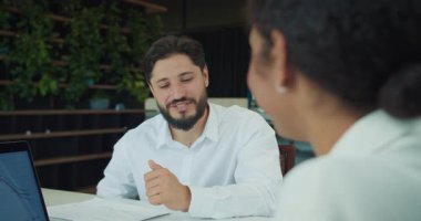 İş adamı, bir meslektaşıyla konuşurken baş parmağıyla gülümsüyor ve jestler yapıyor. Modern bir ofiste tablolar ve bir dizüstü bilgisayar görünür. Yüksek kalite 4k görüntü