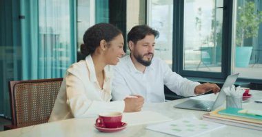 Bir işadamı ve iş kadını modern bir ofiste masa başında oturmuş, bir dizüstü bilgisayarla çalışırken kahve içerek sohbet ediyorlar. Ortam profesyonelce ama rahat. Yüksek kalite 4k görüntü