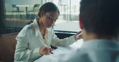 Beyaz bluzlu bir iş kadını, bir meslektaşıyla yaptığı toplantıda basılı grafiği analiz ediyor. Sahne, modern bir ofiste doğal ışık ve profesyonel bir ortamda geçiyor. Yüksek kalite 4k