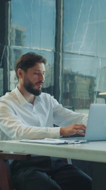 Resmi beyaz gömlekli bir iş adamı, büyük pencereler ve doğal gün ışığıyla çevrili kahve ve belgelerle modern bir ofis masasında dizüstü bilgisayarla çalışıyor..