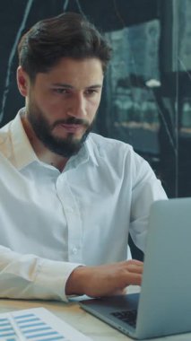 Odaklanmış işadamı modern bir ofiste masa başında oturuyor ve dizüstü bilgisayarla yazı yazıyor. Belgeler ve bir fincan kahve verimli bir iş ortamı olduğunu gösteriyor..