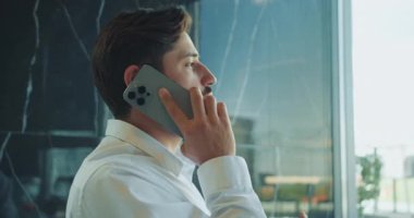 Beyaz gömlekli bir iş adamı modern bir ofiste telefonla konuşuyor. El hareketleriyle bir şeyi açıklamaya çalışıyor gibi görünüyor, iletişimi ve bağlılığı vurguluyor. Yüksek kalite 4k görüntü