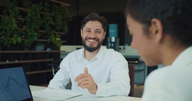 Beyaz gömlekli bir iş adamı modern bir ofiste bir iş arkadaşıyla konuşurken gülümsüyor. Masanın üzerinde dosyalar ve belgeler olan bir dizüstü bilgisayar ekranı var. Yüksek kalite 4k görüntü