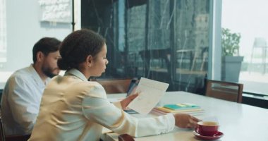 Bir iş kadını, erkek meslektaşı dizüstü bilgisayar üzerinde çalışırken, modern ofis ortamında basılı bir tabloyu inceliyor. Masada takım çalışması olduğunu gösteren belgeler, defterler ve kahve fincanları var. Yüksek kalite 4k