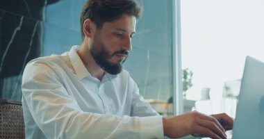 Geniş bir pencereye yakın, parlak ve modern bir ofiste beyaz gömlekli genç bir işadamının dizüstü bilgisayarda yazı yazışının yan görüntüsü. Profesyonel görevlere odaklanmış. Yüksek kalite 4k görüntü