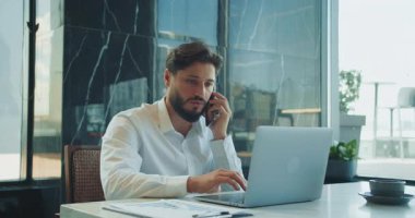 Beyaz gömlekli, kendine güvenen bir işadamı telefonda konuşuyor ve modern bir ofis masasında, parlak bir dizüstü bilgisayarla kahve, belgeler ve doğal ışıkla yazı yazıyor. Yüksek kalite 4k görüntü