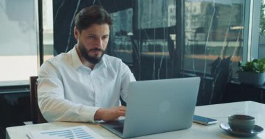 Odaklanmış işadamı modern bir ofiste masa başında oturuyor ve dizüstü bilgisayarla yazı yazıyor. Belgeler ve bir fincan kahve verimli bir iş ortamı olduğunu gösteriyor. Yüksek kalite 4k görüntü