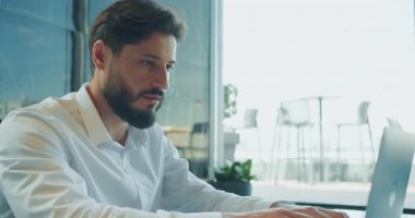 Beyaz gömlekli odaklanmış işadamı, parlak ve modern bir ofiste geniş bir pencerenin önünde otururken dizüstü bilgisayar kullanıyor. Yoğunlaştırılmış ifade verimlilik ve iş akışına işaret ediyor. Yüksek kalite 4k görüntü