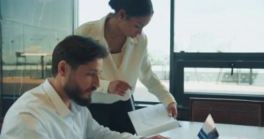 İş kadını, bunu bir masada dizüstü bilgisayarla oturan meslektaşı ile tartışırken basılı bir tabloya işaret ediyor. Modern, güneşli bir ofis alanında birlikte çalışıyorlar. Yüksek kalite 4k görüntü
