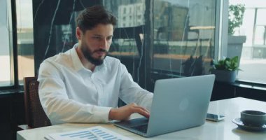 Beyaz gömlekli sakallı bir iş adamı modern bir ofiste dizüstü bilgisayarla çalışıyor. Tablolar ve masadaki kahve fincanı verinin yönlendirdiği bir çalışma oturumuna işaret ediyor. Yüksek kalite 4k görüntü