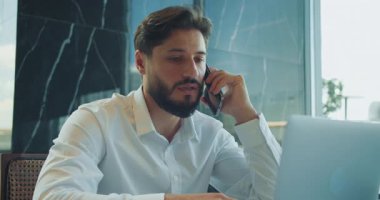 Modern bir ofiste dizüstü bilgisayar üzerinde çalışırken kendine güvenen bir iş adamı telefon görüşmesi yapıyor. Doğal ışık ve çağdaş iç mekanı olan profesyonel bir ortam. Yüksek kalite 4k görüntü