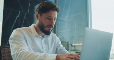 Beyaz gömlekli genç iş adamı, modern bir ofisin penceresinin yanında otururken dizüstü bilgisayarıyla dikkatle çalışıyor. Doğal ışık onun konsantre ifadesini vurgular. Yüksek kalite 4k görüntü