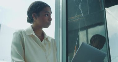 Pencere kenarında dizüstü bilgisayarla çalışan beyaz bluzlu genç bir iş kadını. İfadesi ciddi ve odaklanmış, modern ofis ortamındaki konsantrasyonu vurguluyor. Yüksek kalite 4k görüntü