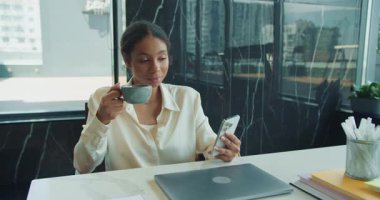 Beyaz bluzlu neşeli bir iş kadını masada akıllı telefonunu kontrol ederken kahvenin tadını çıkarıyor. Ofis malzemeleri ve kapalı bir dizüstü bilgisayar masada görülebilir. Yüksek kalite 4k görüntü