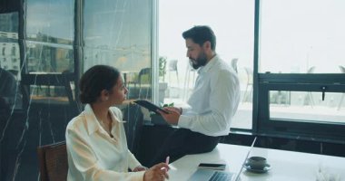 Bir işadamı, elinde tabletle bir masada oturmuş, geniş pencereli modern bir ofiste dizüstü bilgisayar üzerinde çalışan bir iş kadınıyla verileri tartışıyor. Yüksek kalite 4k görüntü