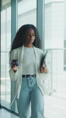 Kendinden emin genç bir kadın, elinde kahve fincanı ve önemli belgelerle enerjik bir ofiste yürüyor.