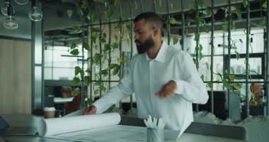 Bitkilerle dolu şık bir ofiste, odaklanmış bir mimar proje geliştirme planlarını dikkatle inceliyor.