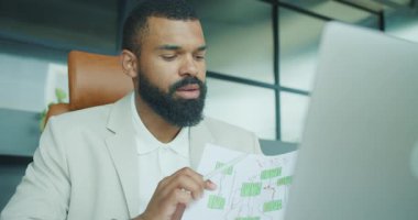 Çalışkan bir işadamı, modern bir ofiste dizüstü bilgisayarı üzerinde çalışırken ayrıntılı çizelge ve raporları gözden geçirir.