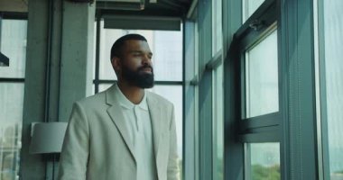 Derin düşüncelere dalmış takım elbiseli bir adam modern ofis alanındaki büyük bir pencereden dikkatlice dışarı bakar.